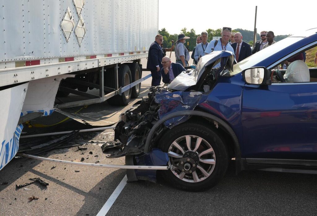 A blue SUV with a crushed front end after crashing into the side of a semi-trailer as several men observe