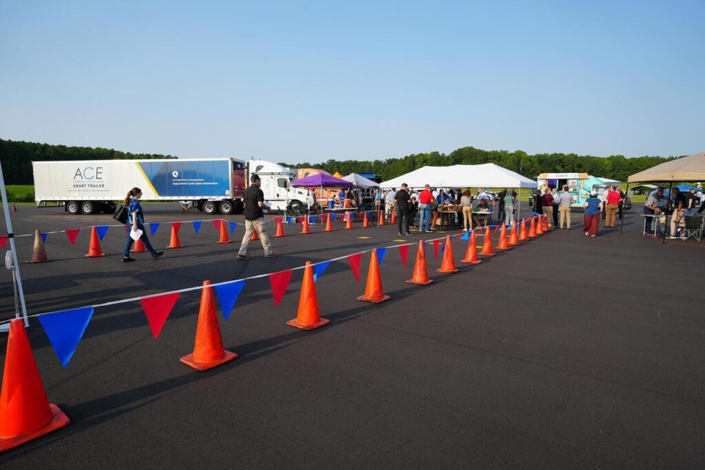 People at an outdoor event with an ACE Smart Trailer and other semi-trucks parked near white tents