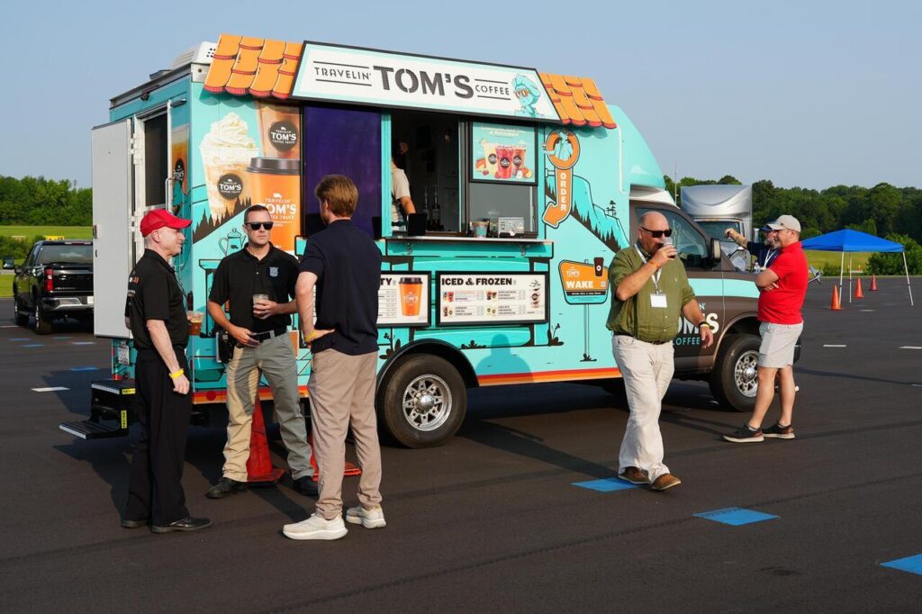 People standing and talking near a Travelin' Tom's Coffee truck parked in an outdoor lot