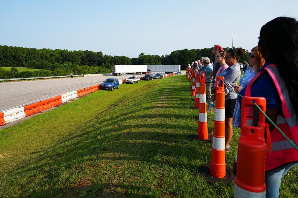 People standing behind traffic barrels watch a vehicle demonstration on a closed road