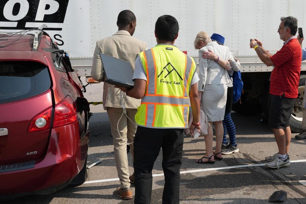 Congresswoman Deborah Ross and Marianne Karth embrace and observers stand near a red car with front-end damage behind a semi-trailer